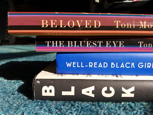 A small stack of the following books: Beloved by Toni Morrison, The Bluest Eye by Toni Morrison, Well-Read Black Girl by Gloria Edem, and Black on a spine with the rest of the title cut off from the picture.